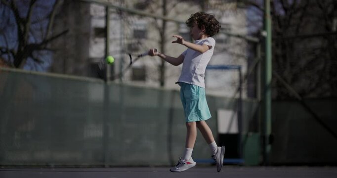 A young boy in a white shirt and blue shorts plays tennis on an outdoor court, showcasing a forehand stroke with determination.