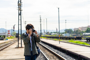 Asian man using digital camera taking picture during walking on railroad track at train station. People enjoy outdoor lifestyle travel countryside by public transportation on summer holiday vacation.