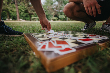 Obraz premium Two people engage in an outdoor board game, using playing cards on a chessboard on a grassy park field. Leisure and strategy combine under the sun.
