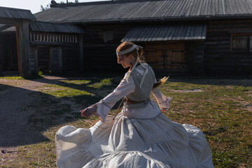 Fototapeta premium A young girl with a long braid in a folk costume is dancing and having fun in a village among wooden architecture, Slavic or Nordic style