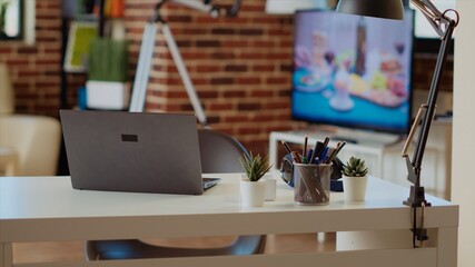 Laptop on desk next to mini plants in empty apartment lit by sunlight, TV left open in background, panning shot. Notebook device in elegant living room with warm interior decor