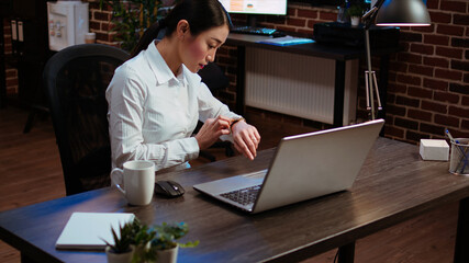 Sleepy employee asleep on computer desk at night in brick wall office, suddenly waking up. Tired businesswoman looking at watch, annoyed by time passing too slow while working overnight, camera A