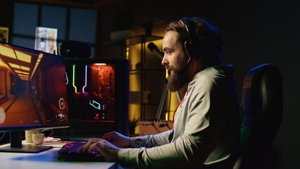 Happy man putting on headphones to communicate with teammates online from his dark living room, playing engaging video games on gaming PC at computer desk, chilling after work © DC Studio