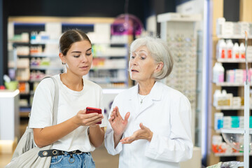 Obraz premium Senior female pharmacist communicates with girl client, looks at mobile phone screen, helps to identify desired drug in photo