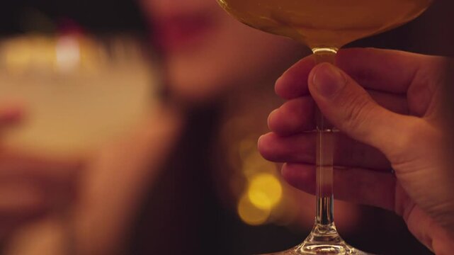 Closeup cropped woman hand making a toast mixed passion fruit smoothie cocktail wearing ring with friend at blurred drink background at nighttime warm light at nightclub counter bar party. Vinosity.