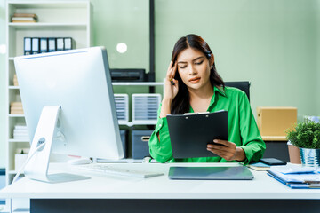 dedicated young Asian businesswoman in a formal suit struggles with stress and office syndrome while working overtime at her desk in a modern glass office. She faces a serious headache and feels sad.