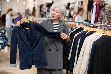 Fototapeta premium Pleased mature woman choosing convenient blazer in clothing shop with large assortment