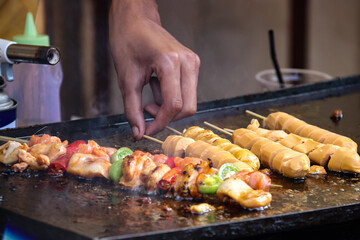  A closeup of a chef grilling sausages and assorted meat skewers with vibrant vegetables. The sizzling meats are being carefully tended to on a hot, oily griddle