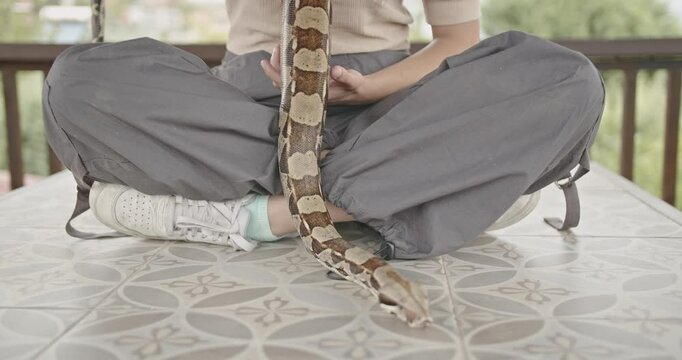 A young woman with pink hair sits peacefully on a tiled patio table, her pet snake draped over her shoulders.