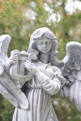 A close-up photo of a white stone statue depicting an angel with wings, playing a harp. The angel has a serene expression and is surrounded by lush greenery.