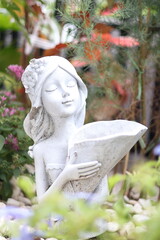 A close-up photo of a white garden statue depicting a woman with closed eyes, holding a seashell in her hands. The statue is adorned with flowers and is surrounded by greenery.