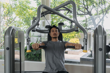Fitness asian Thai man sitting, using chest and shoulder press exercise machine at gym, building...