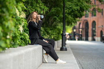Relaxed middle aged woman in business suit sitting on concrete fence among bush plants, drinking...