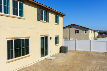 Unfinished backyard view of a new suburban two story house with beige stucco, large energy-efficient windows, sliding glass door and an air conditioning unit bordered by a white vinyl fence