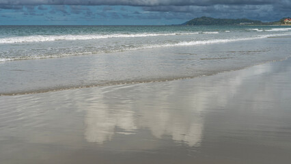 The waves of the turquoise ocean foam and spread over the sandy beach. The blue sky and clouds are reflected on the wet smooth sand. Hills on the horizon. Malaysia. Borneo. Kota Kinabalu.