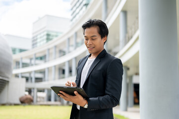 A successful Asian businessman is using his digital tablet while standing in an outdoor hallway.