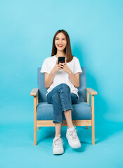 Happy cheerful young woman sitting on the chair and using smartphone on blue background.
