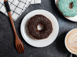 A chocolate-sprinkled donut is placed on a white plate on a dark textured surface. Beside it, a wooden fork rests near a checkered cloth, with a coffee cup and another donut partially visible