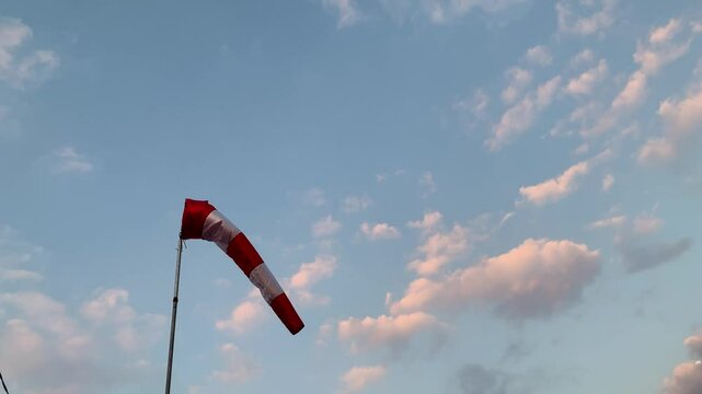 Windsock in the Blue Sky. A red and white windsock against a blue sky with clouds. The windsock is pointing to the left, indicating the direction of the wind.