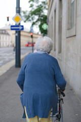 Rear view of elderly woman walking on city street with rollator, going shopping to the store.