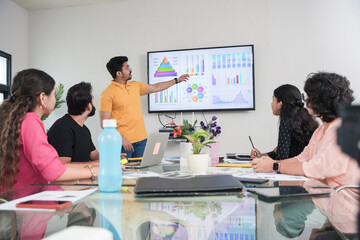 young indian man presents business financial data with charts and graphs on wall tv in the meeting...