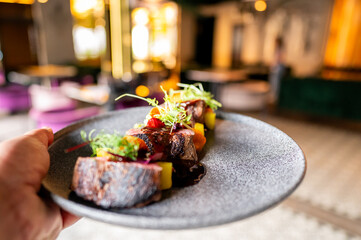A close-up of a hand presenting a gourmet dish on a textured gray plate, featuring succulent pieces of meat, vibrant vegetables, and delicate garnishes, in an elegant restaurant setting.