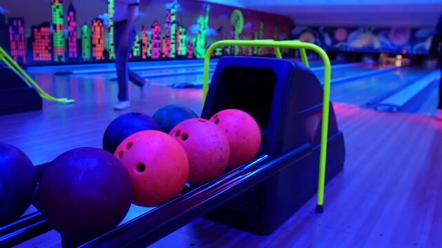 Bowling ball return. Close-up of colorful bowling balls resting in the ball return at a neon-lit bowling alley. A bowler is throwing a ball in the background on another lane