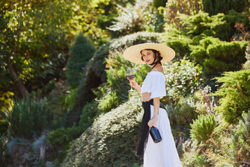 Young woman with wide-brimmed hat, holding glass of red wine, stands in verdant garden, embodying...
