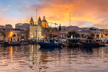 Fototapeta premium Sunset at Marsaxlokk, Malta picturesque fishing village, with colorful traditional boats reflecting in tranquil waters.