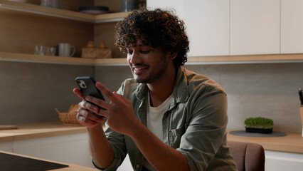 Satisfied Indian male win with phone winning financial prize reward goal achievement with mobile app betting service online victory. Arabian happy man celebrating smartphone success at home kitchen