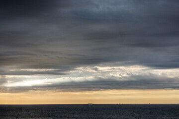 A dramatic panoramic ocean view under stormy skies during sunset, featuring dark clouds and distant ships on the water