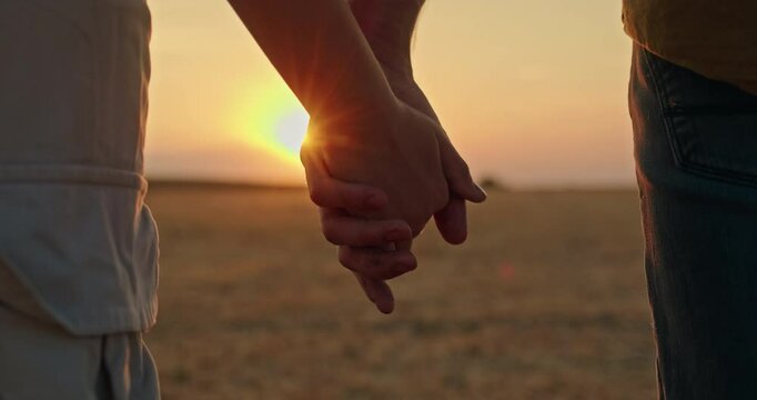 The hands of a farmer couple are illuminated by the setting sun as they hold hands in front of a field where a combine harvester works in the distance.