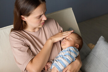 Upset woman measuring her child's temperature with her hand worried about the infants illness checking her baby's forehead for signs of fever taking care of the sick infant at home