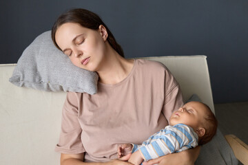 Stressed tired mom taking care of her infant child and feeling fatigue sitting in sofa leaning on soft pillow keeping eyes closed needs rest while taking care of her newborn alone