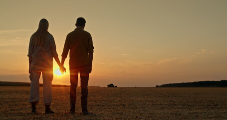 A farmer couple holds hands as they admire the sunset over a field with a combine harvester working in the distance, symbolizing the family farm concept.