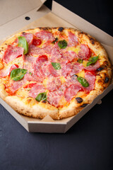 Pizza with cheese, ham, green basil leaves and tomatoes in paper box on black background. Food product advertising banner, copy space