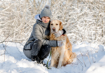 Teenage Girl And Golden Retriever Sit Together In Snow-Covered Forest During Winter