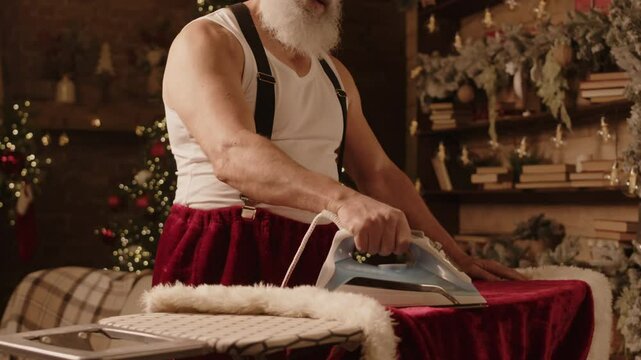 Medium shot of positive elderly Caucasian gentleman singing and enjoying festive dance while ironing Santa Claus costume getting ready for Christmas celebration