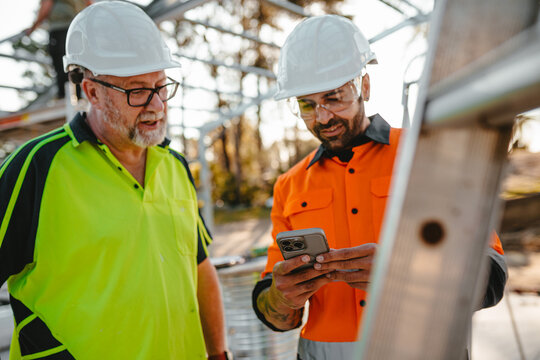 Two men wearing safety gears on the site with one holding a mobile device focus on phone