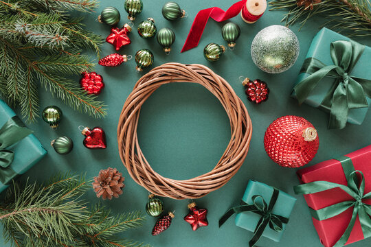 Overhead view of a wicker wreath surrounded by wrapped Christmas gifts, fir branches, Christmas baubles, ornaments, pinecones and ribbon on a green background - Powered by Adobe