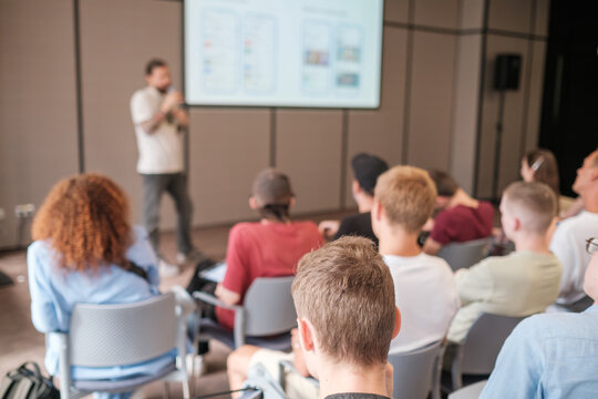 Group of people attending seminar, focusing on speaker presenting at front of conference room with projector screen.