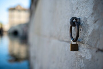 A small lock on an old metal mooring ring. Stone wall on a riverside in Europe, shallow depth of field, river water and building in the background, no people