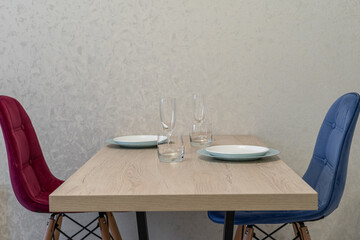 the design of a modern small kitchen in the apartment. There are plates and glasses on the wooden table, and red and blue upholstered chairs near the table.  Setting the table for breakfast.