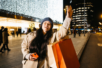 Young Asian woman waving to catch a taxi on a street at night, holding shopping bags after a...