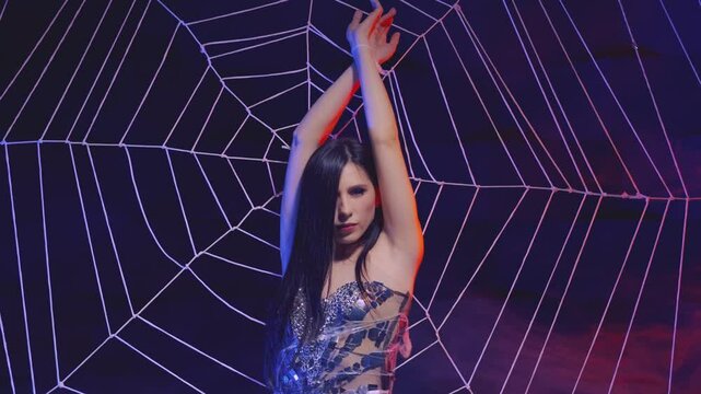 sexy woman victim of spider hanging tied on huge web. fantasy girl gothic dark queen in evening dress. dark studio creative scenery design. performance show night club celebration party halloween