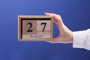Female Hand holding wooden block calendar with the date August 27 on a blue background