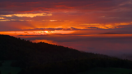 LENS FLARE, SILHOUETTE: Dramatic sunset and burning sky over forested hilly landscape. The clouds are ablaze with warm and vivid colors as the setting sun peeks through them with its golden rays.