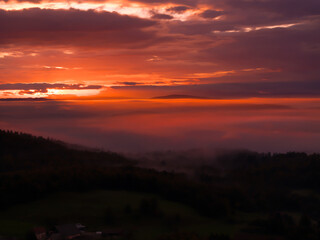 The beautiful colors of an autumn sunrise, glowing over a valley hidden in mist. An ethereal view of a hilly landscape with vibrant colors lighting up the cloudy morning sky as night turns to day.