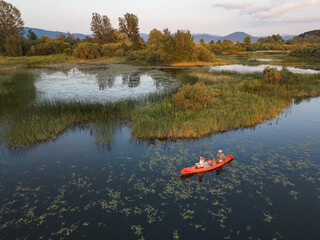 Fototapeta premium Couple enjoying romantic traveling with a canoe on the calm lake during summer sunset, aerial shot. Outdoor wellness concept.
