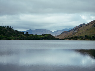Serene lake surrounded by rolling hills and mountains under an overcast sky at midday